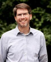 Ben Carson with short brown hair graying at the temples, brown and gray mustache and beard wearing glasses and a light blue button down shirt