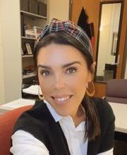 Dr. Ashley Rodrigues smiling with straight brown hair pulled back with a headband wearing a black knit vest over a white button-down blouse