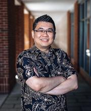 Dr. Cory Yeo smiling with short spiked black and gray hair wearing black rim glasses and a black short sleeve button down shirt with tropical leaves outlined in beige