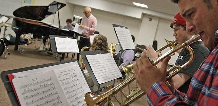 Jazz band class with students playing horns in the foreground, students playing saxophones, a professor, a piano and drum set in the background.  