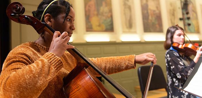 a BSU student in foreground playing the cello and another student in the background playing violin